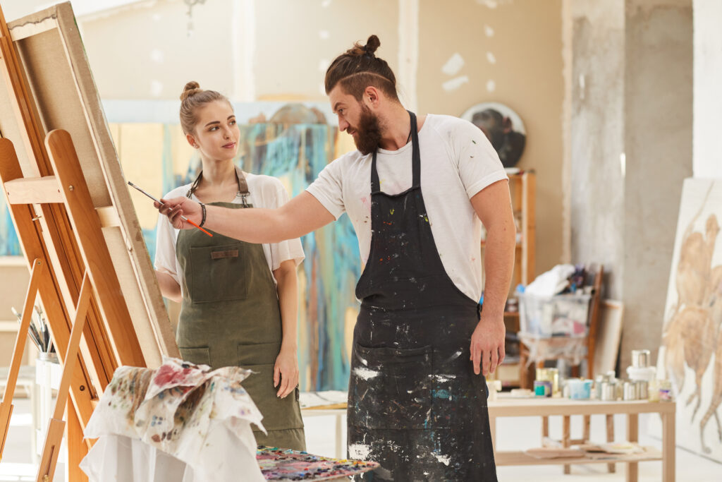Portrait of contemporary couple painting picture together while working in art studio, focus on bearded man wearing apron and holding paintbrush, copy space