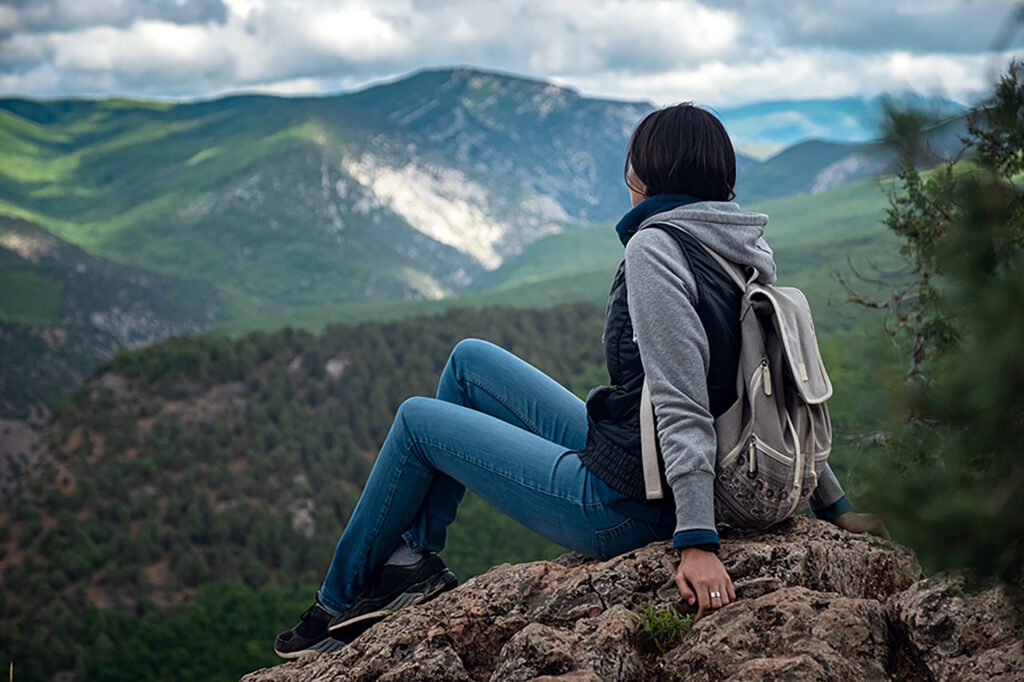 Young traveling woman with a backpack and sitting on top of the mountain cliff with relaxing mood and watching beautiful view of woods and blue sky and clouds on vacation. Asian woman travel alone.